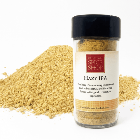 A clear plastic spice jar with a black lid labeled 'Old Town Spice Shop Hazy IPA' stands in front of a mound of the same light tan seasoning powder on a white background.