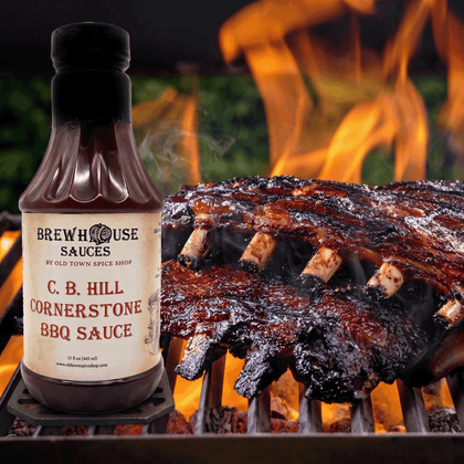 Bottle of C. B. Hill Cornerstone BBQ Sauce next to ribs on a grill with flames in the background