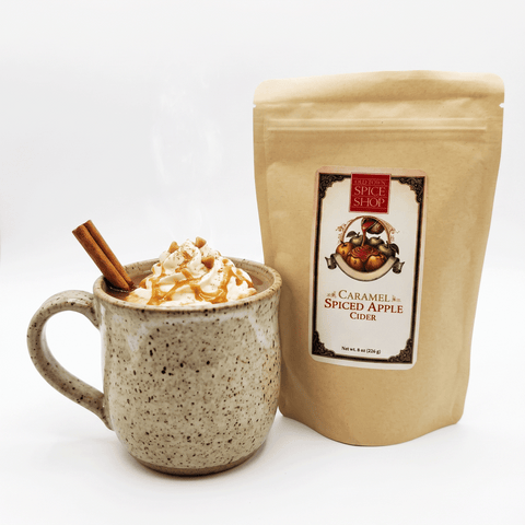A kraft paper pouch of Spiced Apple Cider by Spiced Spice Store stands next to a speckled ceramic mug filled with whipped cream, caramel drizzle, and a cinnamon stick, on a clean white background.