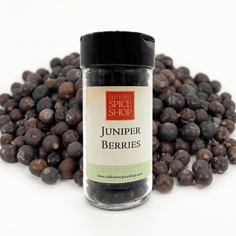 A bottle of Old Town Spice Shop Juniper Berries centered in front of a pile of dark purple-black juniper berries on a white background.