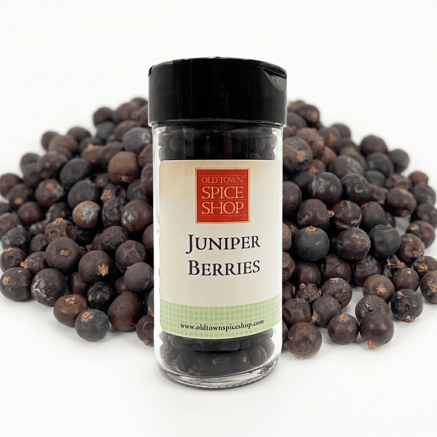 A bottle of Old Town Spice Shop Juniper Berries centered in front of a pile of dark purple-black juniper berries on a white background.