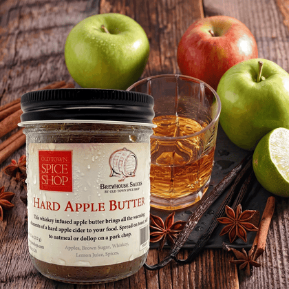 Jar of Old Town Spice Shop Hard Apple Butter with a glass of whiskey, apples, and spices on a wooden surface.