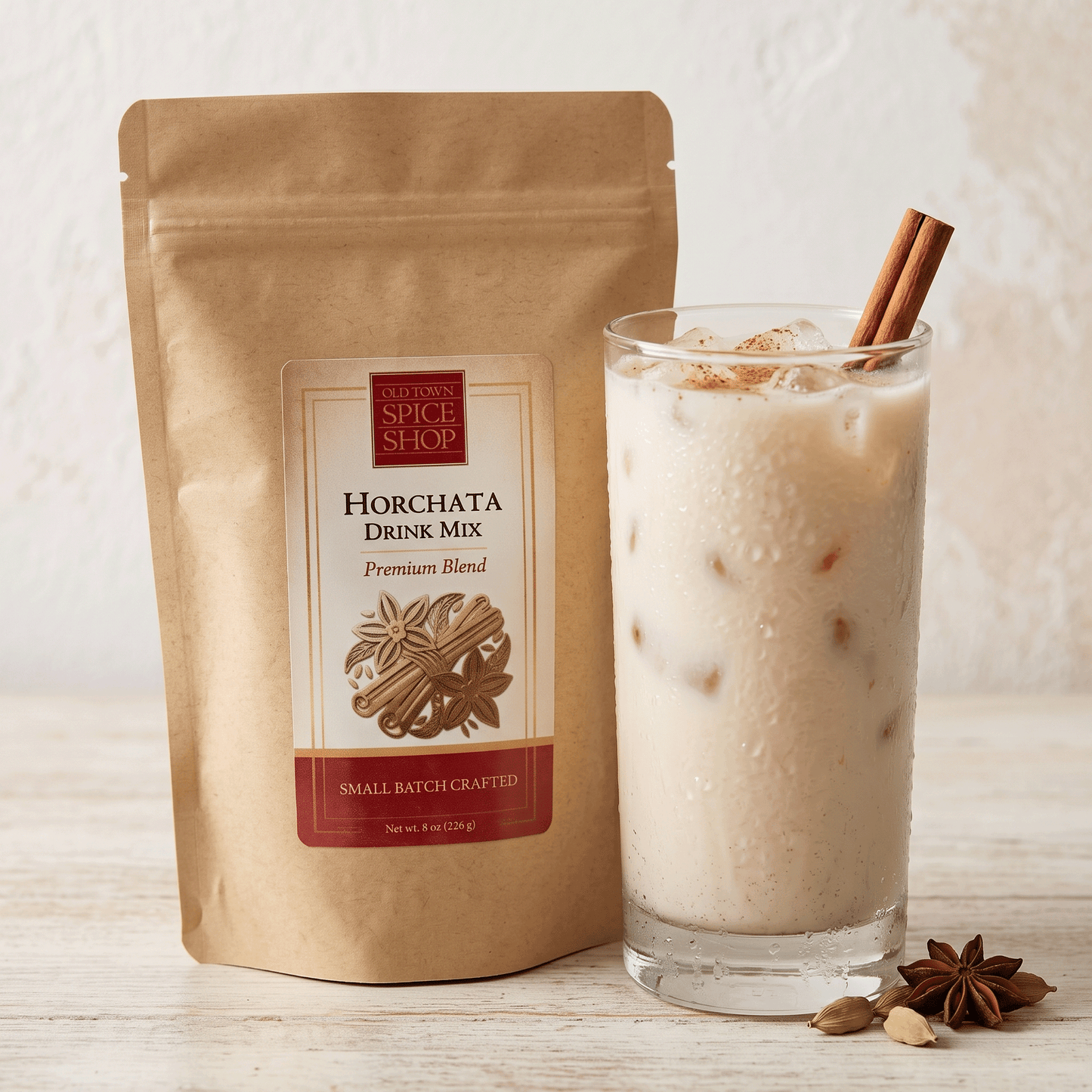 A bag of  Horchata Drink Mix next to a glass of iced horchata with cinnamon sticks, on a light wood surface against a textured white wall.