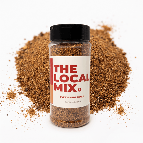 A clear plastic shaker bottle of 'The Local Mix Everything Shake' with a black lid and white label, set against a white background with a pile of brown spice mix behind it.