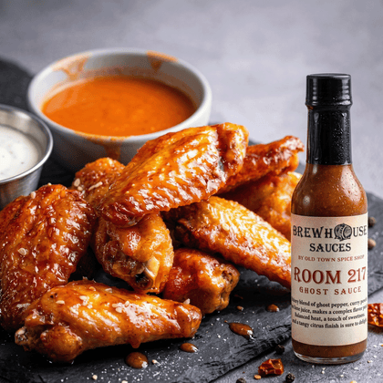 Baked chicken wings with a bottle of Brewhouse Sauces Room 217 Ghost Sauce on a dark surface.