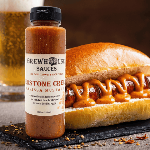 Bottle of Brewhouse Sauces Redstone Crema Harissa Mustard with a hot dog on a bun being drizzled with the sauce.