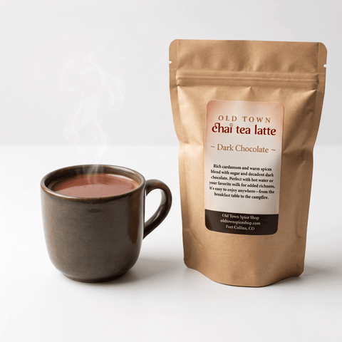A kraft paper stand-up pouch of Old Town Chai Tea Latte Dark Chocolate next to a dark ceramic mug filled with steaming chai tea, on a clean white background.