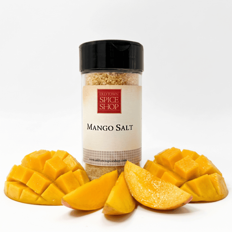 A clear spice jar labeled 'Old Town Spice Shop Mango Salt' with a black lid, centered on a white background and surrounded by fresh mango slices and diced mango pieces.