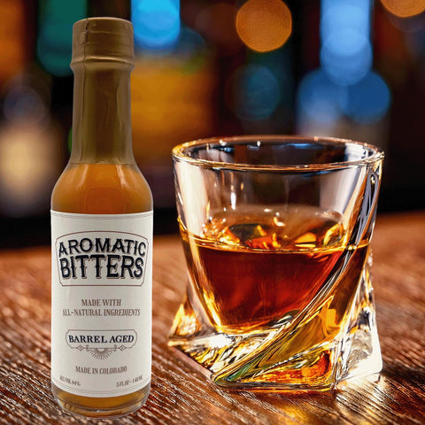 A bottle of Aromatic Barrel-Aged Bitters next to a glass of whiskey on the rocks, with a dimly lit bar in the background.
